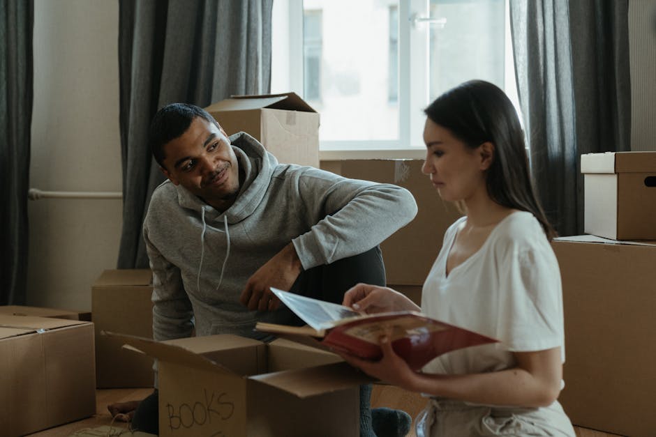 Inside a room with a large window and grey curtains, a man with dark skin, wearing a grey hoodie, is crouched down next to a woman with light skin and long dark hair, dressed in a white top. They are surrounded by moving boxes made of cardboard, some labeled with black marker, such as one marked 'BOOKS', and others stacked on the floor and shelf. The woman is holding an open book or folder and looking at it, while the man is looking attentively at her, suggesting a discussion about the house move or packing process. Behind them, a cardboard box is partially opened, and in the background, a cardboard box and a large window with daylight illuminating the scene are visible, indicating an active home relocation or moving situation. The environment reflects a typical packing and moving phase managed by Man with Van Harrow, involving furniture, packaging materials, and the logistics of a home transfer.