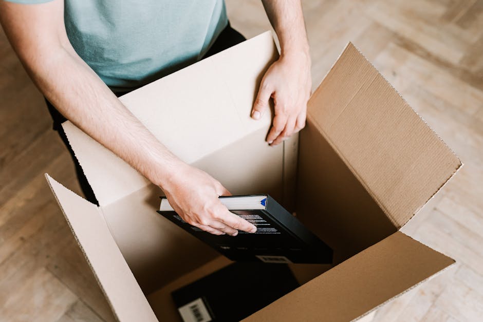 A person with visible arms, dressed in a light blue shirt, is placing a black boxed electronic device into a large, open cardboard box on a wooden floor. The cardboard box, with flaps folded outward, already contains other packing materials, including a cardboard container and bubble wrap. Nearby, a white plastic wrapped item and additional cardboard pieces can be seen. The image captures a packing process typically associated with house removals or home relocation services, illustrating careful handling and packing of belongings, which is relevant to furniture transport and moving logistics managed by [COMPANY_NAME], such as [PAGE_TITLE]. The setting appears to be inside a property, emphasizing the preparation phase of a residential move, with natural lighting illuminating the scene to focus on the packing activity involved in furniture transport and packing and moving tasks.