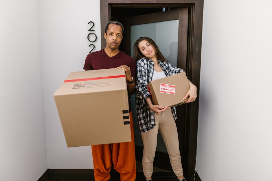 A man and a woman standing inside an entryway, positioned just outside a doorway with a dark wooden frame, are holding cardboard boxes used for home relocation. The man, wearing an orange pair of trousers and a maroon t-shirt, holds a large cardboard box with a red tape seal. The woman, dressed in light-colored pants and a checked shirt, holds a smaller cardboard box labeled 'Fragile.' The background includes a white wall with black letters spelling '200,' indicating the apartment or unit number, and the lighting is bright, highlighting the details of packaging materials such as cardboard and tape. The scene captures the loading process involved in furniture transport and packing for a house move, consistent with professional removals services carried out by [COMPANY_NAME], demonstrating the careful handling of items during a home relocation.