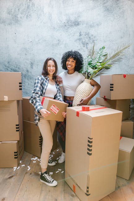Two women are standing inside a room filled with cardboard moving boxes, which vary in size and are sealed with red tape. One woman, on the left, is wearing a plaid shirt over a white top and beige pants, holding a small cardboard box in her hands. The other woman, on the right, is dressed in a white top and dark checkered trousers, holding a large woven basket with green houseplants that include long, slender leaves and broader, textured foliage. The background features a textured light grey wall, and the floor is light wood with scattered packaging remnants. The scene captures a moment during home relocation, with the women preparing items for transport, possibly as part of a furniture transport or packing process managed by [COMPANY_NAME], specialising in removals and moving services. The setting and objects illustrate the packing and moving activities involved in a house move, reflecting the professional handling of household items for moving day.