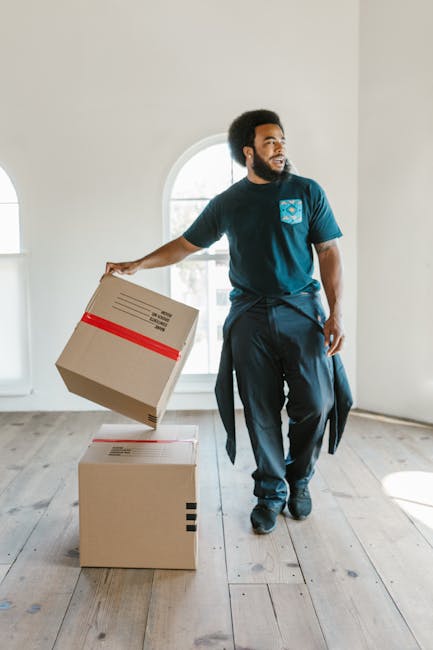 A man and a woman standing inside an entryway, positioned just outside a doorway with a dark wooden frame, are holding cardboard boxes used for home relocation. The man, wearing an orange pair of trousers and a maroon t-shirt, holds a large cardboard box with a red tape seal. The woman, dressed in light-colored pants and a checked shirt, holds a smaller cardboard box labeled 'Fragile.' The background includes a white wall with black letters spelling '200,' indicating the apartment or unit number, and the lighting is bright, highlighting the details of packaging materials such as cardboard and tape. The scene captures the loading process involved in furniture transport and packing for a house move, consistent with professional removals services carried out by [COMPANY_NAME], demonstrating the careful handling of items during a home relocation.