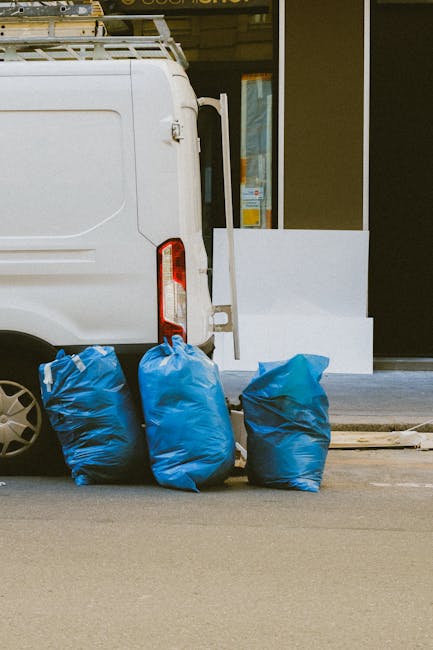Close-up view of the rear section of a white commercial van parked on a city street, with three large blue plastic garbage bags placed in front of it, likely containing household waste or packing materials. The van is positioned near a building with a glass window and a partially visible entrance, indicating an ongoing home relocation or removal process. The rear doors of the van are closed, and the vehicle's rear light is visible. The setting suggests preparation for waste disposal or clearance work associated with furniture transport or packing during house removals. The image emphasizes the logistics involved in moving services offered by Man with Van Harrow, particularly in managing waste collection and loading for residential moves, with a focus on efficient clearance of packing materials and unwanted items as part of a furniture transport or home relocation service.