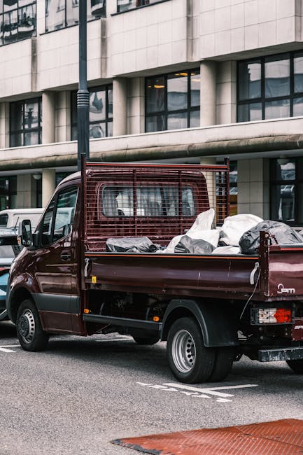 A dark red, small flatbed van parked on a city street in front of a modern building with large windows. The van's cargo area is filled with various household items, including cardboard boxes, plastic bags, and wrapped furniture, indicating a home relocation or moving process. The truck has a metal mesh partition behind the cab for safety, and there are a few other vehicles parked nearby. The loading area shows no specific moving equipment in use, but the presence of packed belongings suggests recent or ongoing packing and loading activities. Man with Van Harrow, a company specializing in removals, often manages such home transport tasks, including packing, loading, and transportation, as part of their comprehensive moving services.