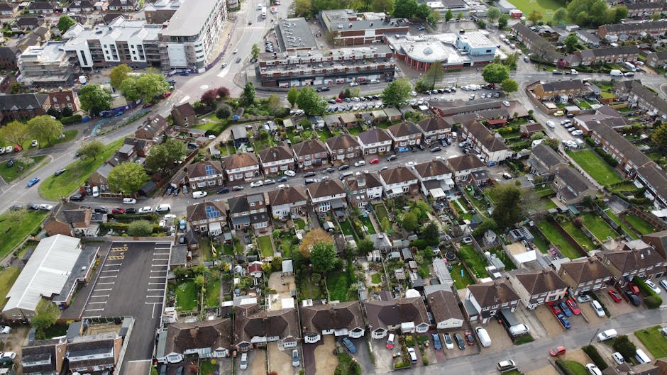An aerial view of a residential neighbourhood showing a row of terraced houses with small gardens and driveways, parked cars along the streets, and a large commercial building at the top of the image. In the foreground, a house with a green lawn and a backyard contains outdoor furniture and garden features. A moving van from Man with Van Harrow is parked on the street near this property, with visible furniture and boxes being carefully loaded or unloaded during a home relocation. The surrounding area includes trees, paved pathways, and additional residential buildings, all captured in natural daylight. The scene illustrates the logistics involved in furniture transport and packing and moving services, addressing challenges like tight parking, typical of house removals in Harrow.