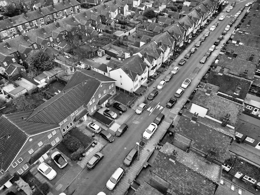 Aerial black-and-white photograph of a residential street in Harrow showing rows of terraced houses with tiled roofs, parked cars lining both sides of the narrow road, and a designated loading zone near a house where Man with Van Harrow is performing a home relocation. The scene includes a parked van on the pavement, partially blocking a driveway, with furniture and packed boxes visible inside and around the vehicle. The street is tightly packed with vehicles and houses, illustrating the challenge of tight parking during a furniture transport and moving process, as part of a house removal service. The image captures the detailed environment including some trees, utility poles, and small front gardens, conveying the logistical complexities of loading and unloading household items in a residential setting.
