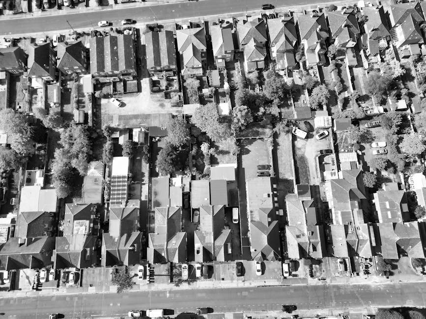 An aerial view of a residential neighbourhood showing a row of terraced houses with small gardens and driveways, parked cars along the streets, and a large commercial building at the top of the image. In the foreground, a house with a green lawn and a backyard contains outdoor furniture and garden features. A moving van from Man with Van Harrow is parked on the street near this property, with visible furniture and boxes being carefully loaded or unloaded during a home relocation. The surrounding area includes trees, paved pathways, and additional residential buildings, all captured in natural daylight. The scene illustrates the logistics involved in furniture transport and packing and moving services, addressing challenges like tight parking, typical of house removals in Harrow.
