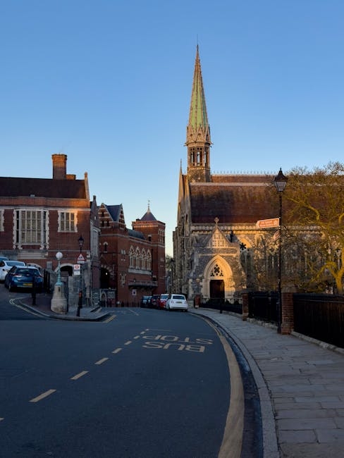 A narrow street in Harrow with a loading area for house removals in front of a historic church featuring a tall, pointed green spire and arched entrance. The street is paved with asphalt and has yellow dashed lines and bus stop markings. On the street, several parked cars are visible, with some near the curb and others further along the road. To the right, a sidewalk runs parallel to the street, separated by a low black metal fence and lined with a streetlamp. The scene is illuminated by natural daylight, emphasizing the brick and stone facades of the surrounding buildings and the church. This setting demonstrates the typical environment for furniture transport and packing during a house relocation in Harrow, with visible elements such as vehicles, packaging materials, and loading space that relate to professional removals services like those from Man with Van Harrow.