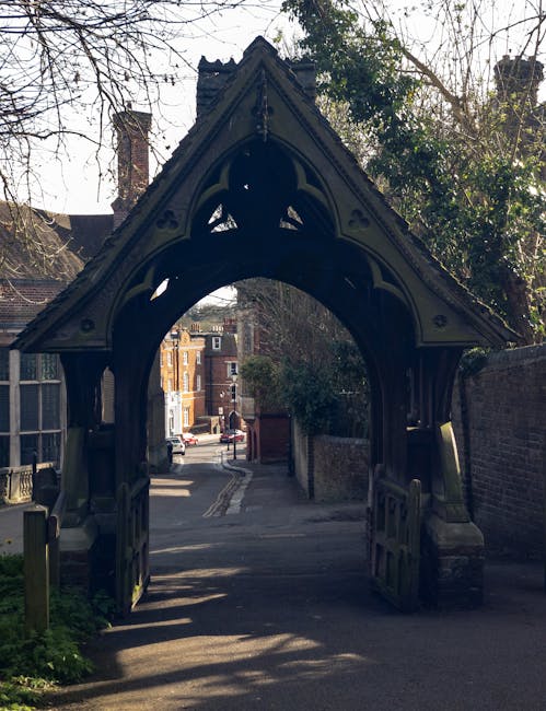 A historic, ornate wooden gateway with decorative carvings and a pointed arch overlooks a narrow street in a residential area. The gateway is situated at the entrance to a private property, with a brick wall on one side and trees in the background. Sunlight casts shadows on the pavement and the surrounding buildings, which include traditional brick houses with chimneys visible further down the street. The scene captures the quiet atmosphere of a local neighbourhood, with no vehicles or people in the immediate vicinity. This type of structure is commonly seen in historic parts of Harrow, where house removals and packing services by companies such as Man with Van Harrow facilitate home relocations, including navigating narrow streets like this one during furniture transport and loading processes.