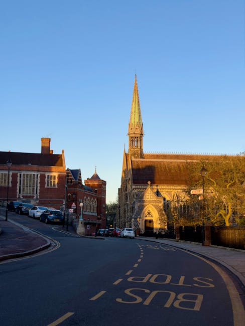 A narrow street in Harrow with a loading area for house removals in front of a historic church featuring a tall, pointed green spire and arched entrance. The street is paved with asphalt and has yellow dashed lines and bus stop markings. On the street, several parked cars are visible, with some near the curb and others further along the road. To the right, a sidewalk runs parallel to the street, separated by a low black metal fence and lined with a streetlamp. The scene is illuminated by natural daylight, emphasizing the brick and stone facades of the surrounding buildings and the church. This setting demonstrates the typical environment for furniture transport and packing during a house relocation in Harrow, with visible elements such as vehicles, packaging materials, and loading space that relate to professional removals services like those from Man with Van Harrow.