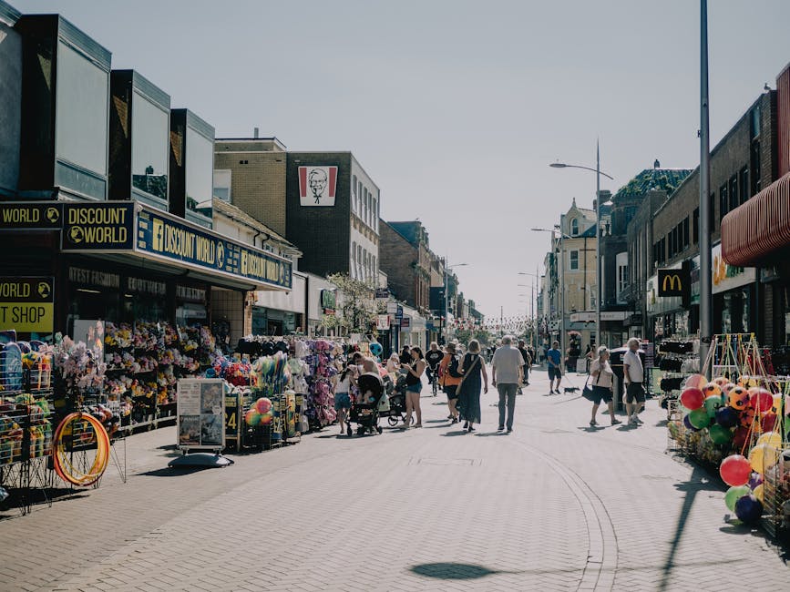 A street scene in Harrow town centre showing pedestrians walking along a busy commercial area with various shops on both sides. On the left side, there are storefronts including a discount shop with outdoor displays of colorful plastic balls, balloons, and toys, along with wire baskets and packaging materials. The shops display signs, and the pavement is paved with light-colored bricks. The street is lined with buildings of brick, glass, and concrete, with other retail outlets visible, including one with a McDonald's logo on the right side. The scene is illuminated by daylight with clear weather, and there are street lamps and signage overhead. The image captures ongoing activity typical of a shopping district, providing context for local relocation or moving services such as furniture transport, packing, and loading activities that Man with Van Harrow might undertake in similar environments.