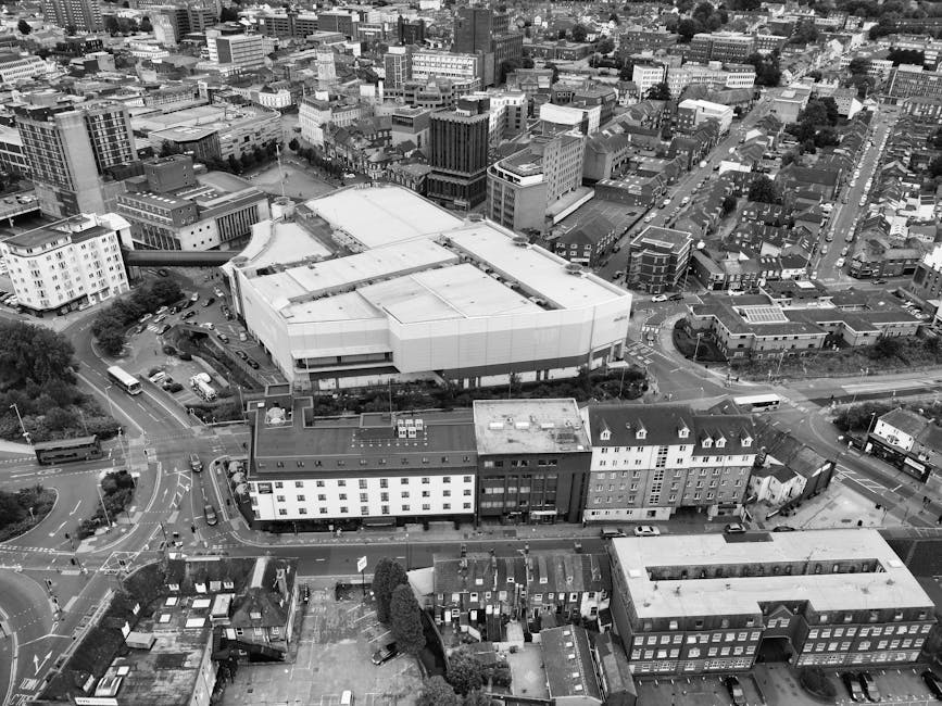 An aerial view of a dense urban area showing a large, modern building with a flat roof, surrounded by smaller multi-storey structures and residential buildings. The scene includes roads with visible vehicles, including buses and cars, navigating around the buildings and intersections. In the foreground, there is a parking lot with several parked cars and trees providing some greenery. The photograph captures the cityscape during daylight, with the overall environment indicating active urban area planning and development. This setting relates to house removals and moving services, as it highlights an urban context where relocations and furniture transport may occur, similar to the services offered by Man with Van Harrow during office removals or home relocations in Harrow town centre.
