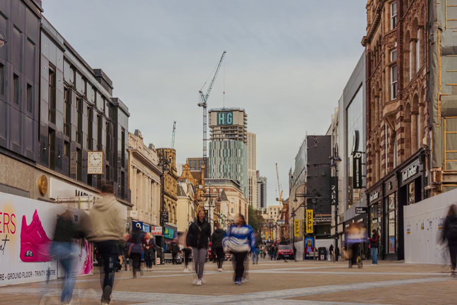 A street scene in Harrow town centre showing pedestrians walking along a busy commercial area with various shops on both sides. On the left side, there are storefronts including a discount shop with outdoor displays of colorful plastic balls, balloons, and toys, along with wire baskets and packaging materials. The shops display signs, and the pavement is paved with light-colored bricks. The street is lined with buildings of brick, glass, and concrete, with other retail outlets visible, including one with a McDonald's logo on the right side. The scene is illuminated by daylight with clear weather, and there are street lamps and signage overhead. The image captures ongoing activity typical of a shopping district, providing context for local relocation or moving services such as furniture transport, packing, and loading activities that Man with Van Harrow might undertake in similar environments.