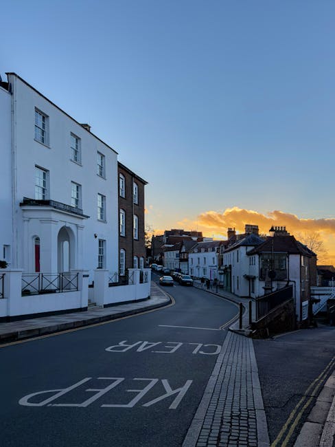 A quiet street scene during sunset featuring a mix of modern and traditional terraced houses, with some buildings painted white and others in darker shades, situated along a gently curved road. The ro