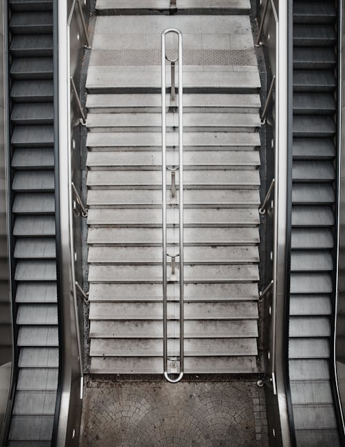 A top-down view of a staircase within a building, showing grey concrete steps with a central metal handrail running along the middle. The staircase is flanked by two sets of black and grey metal escalators, with the left side visible and slightly blurred. The surrounding area includes grey tiled flooring at the top of the stairs and a patterned curved floor surface at the base. The lighting is bright and evenly distributed, highlighting the clean, well-maintained surfaces. This staircase area is part of a premises where home relocation or furniture transport might occur, and the image depicts the access point involving stairs that [COMPANY_NAME] may need to navigate during a house removal, as detailed on the page about stairs and lift access in South Harrow.