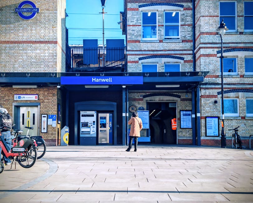 Outside Hanwell underground station entrance, a woman wearing a beige coat and dark trousers is standing near the ticket machines and information boards, with her back facing the camera. To her left, 